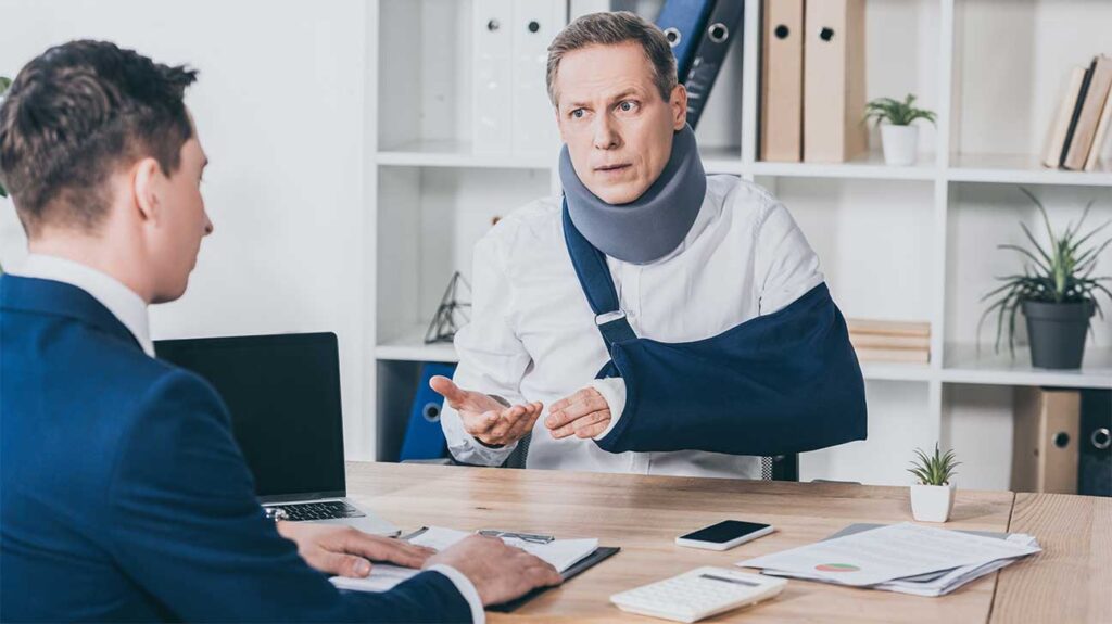 Injured man with a neck brace and arm sling speaks with a professional at a desk